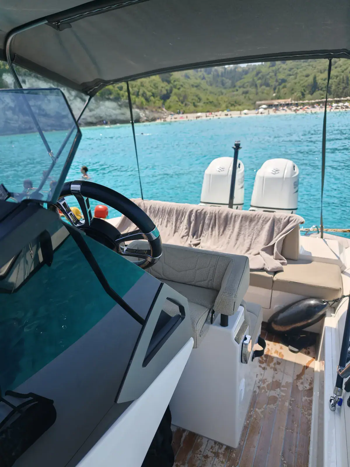 The cockpit of Emerald from the bow side: the helm seat and wheel, the rear bench with towels over it, the teak sole and the stern, with turquoise water and a wooded shore beyond