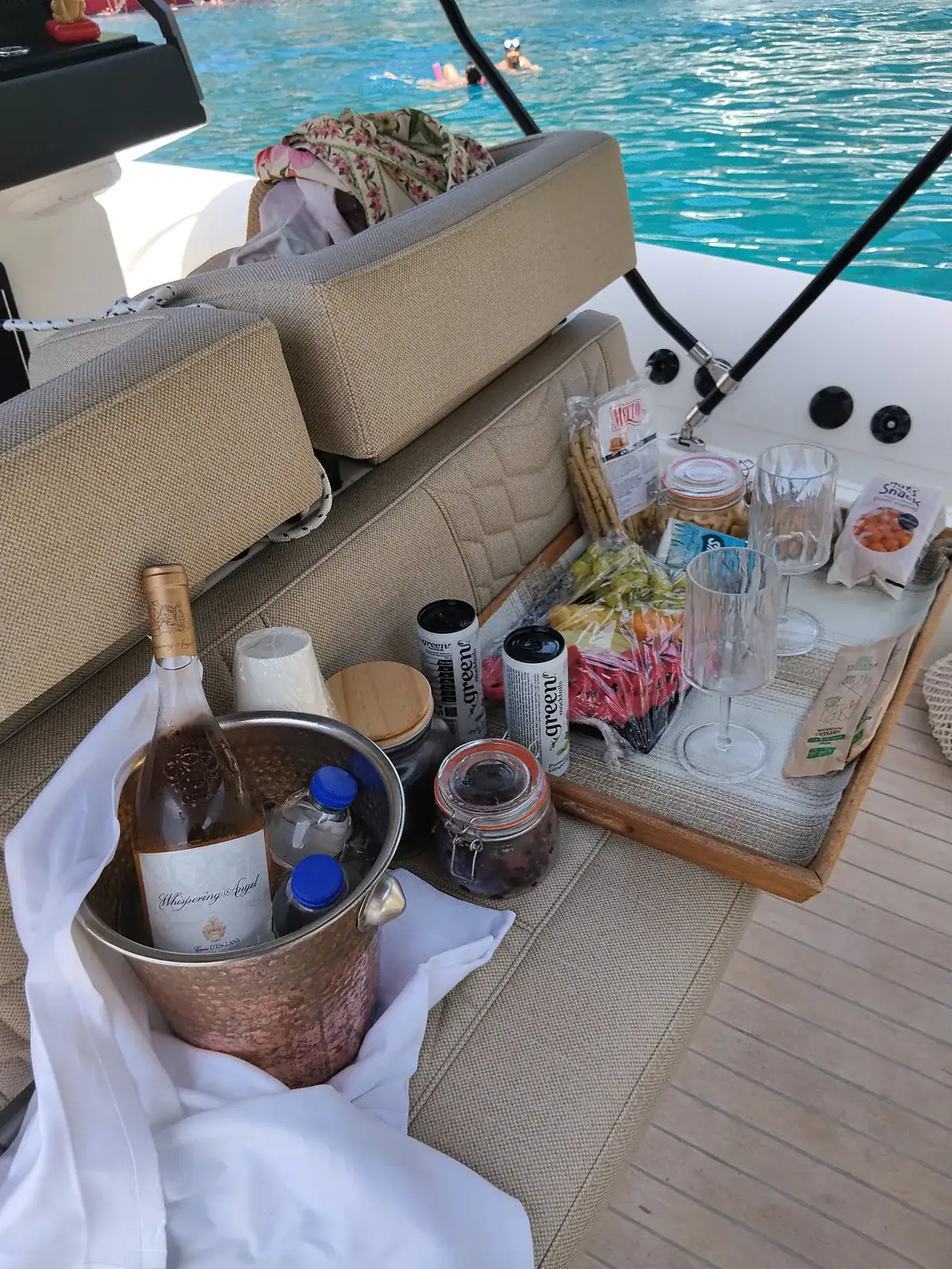 A tray of water, soft drinks, wine, fruit and snacks set out on the aft seat, with a swimmer in turquoise water beyond