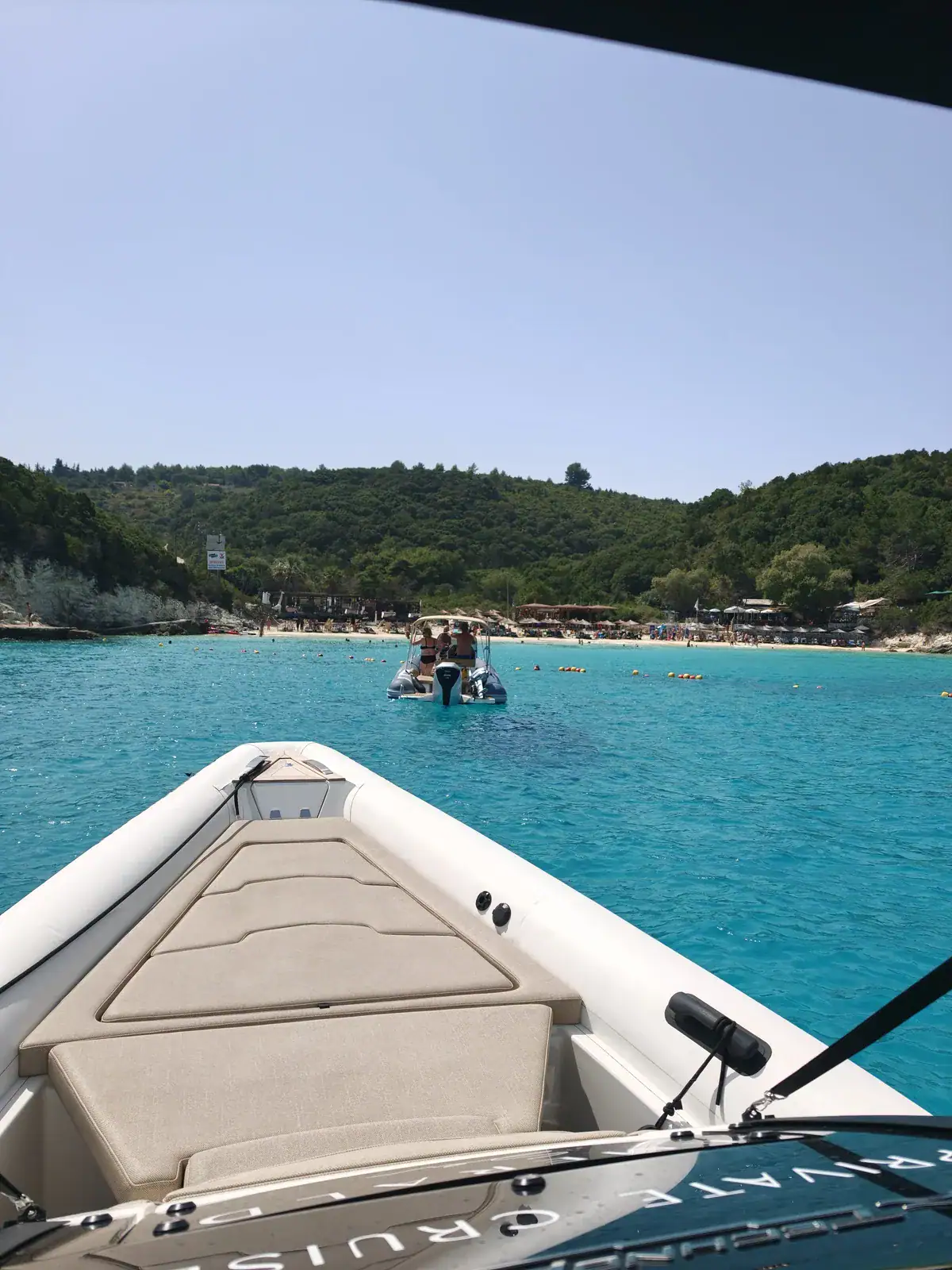 The view forward from beneath Emerald’s canopy: the dark canopy edge across the top of the frame, the pale padded sunpad across the bow below it, turquoise water and a wooded bay beyond